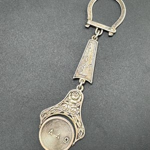 May include: A silver-toned metal keychain featuring a circular ring at the top, a central trapezoidal pendant engraved with the text KPH and a wing motif, and a lower section decorated with floral rose carvings holding a spinning circular disc with AAO.