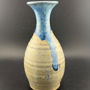 以下が含まれることがあります： 青とベージュの配色が施された手作りのセラミック花瓶。花瓶は、フレアの青いリムと側面に青いドリップデザインが施されています。花瓶の本体は、水平のテクスチャラインが入ったベージュ色です。