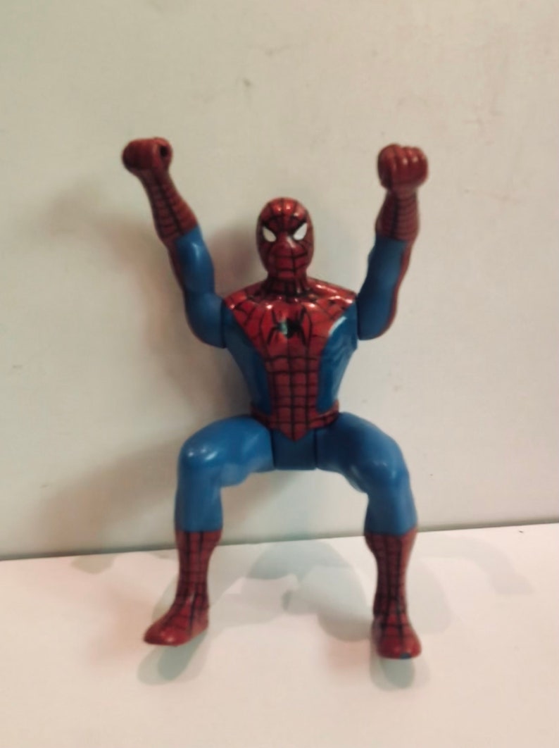 May include: A red and blue plastic action figure of Spider-Man with his arms raised in the air. The figure is posed in a dynamic action pose.