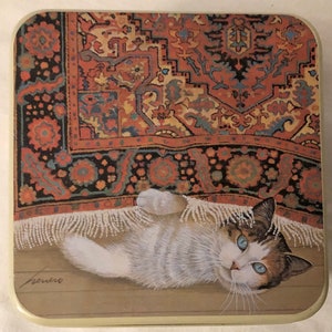 May include: A white cat with blue eyes lies on its back under a colorful rug with fringe. The cat is looking up at the viewer. The rug has a traditional Persian design.