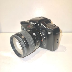 Könnte beinhalten: Eine schwarze Minolta Maxxum 7xi Filmkamera mit einem Minolta-Objektiv. Die Kamera hat ein silbernes Etikett mit der Aufschrift "Minolta Maxxum 7xi".