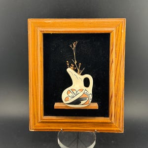 Puede incluir: Una pieza decorativa enmarcada con una pequeña jarra de cerámica color crema con un diseño negro, naranja y blanco. La jarra contiene flores secas y se encuentra sobre una repisa de madera sobre un fondo negro, dentro de un marco de madera.