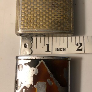 May include: A vintage silver and gold lighter with a patterned design. The lighter is 1 inch tall and has a brown and silver bottom with a silver design. The lighter is in used condition and has some wear and tear.
