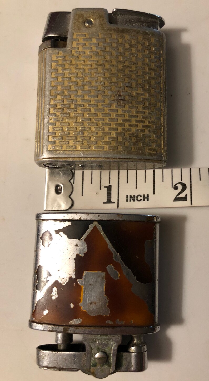 May include: A vintage silver and gold lighter with a patterned design. The lighter is 1 inch tall and has a brown and silver bottom with a silver design. The lighter is in used condition and has some wear and tear.