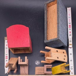 May include: A collection of miniature wooden furniture pieces, including chairs, a red cabinet, and a gray upholstered box. The pieces are in various states of completion and are arranged on a dark surface. A ruler is placed next to the objects for scale.
