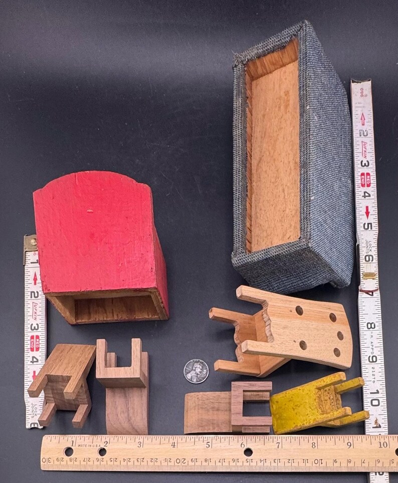 May include: A collection of miniature wooden furniture pieces, including chairs, a red cabinet, and a gray upholstered box. The pieces are in various states of completion and are arranged on a dark surface. A ruler is placed next to the objects for scale.