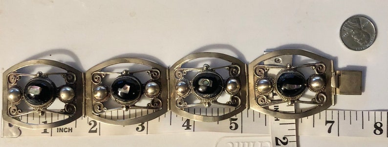 Puede incluir: Pulsera vintage plateada con cuatro eslabones rectangulares. Cada eslab&oacute;n presenta una piedra negra ovalada central, flanqueada por esferas plateadas y filigranas decorativas. La pulsera mide aproximadamente 18 cm de largo.