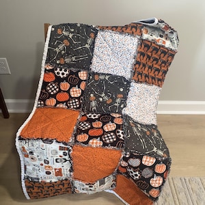 May include: A patchwork rag quilt featuring a variety of Halloween-themed fabric squares. The patterns include skeletons, orange and black pumpkins, black cats, and various holiday icons on white, orange, grey, and black backgrounds.