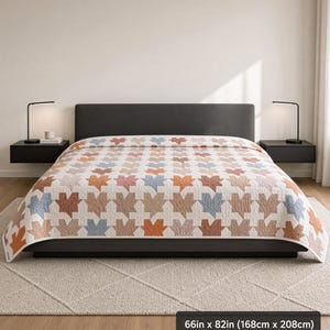 Könnte beinhalten: Eine Steppdecke mit den Maßen 168 cm x 208 cm mit einem sich wiederholenden geometrischen Muster aus stilisierten Blättern in Terrakotta, Staubblau und Beige auf weißem Grund, drapiert über einem dunkelgrauen Bettrahmen mit passendem Kopfteil.
