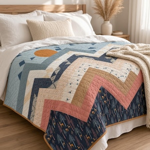May include: A quilt featuring a geometric chevron pattern in shades of navy blue, dusty pink, tan, and cream. The design includes printed motifs of deer and pine trees, a solid orange sun, and small blue diamonds. The quilt has a thin mustard yellow border.