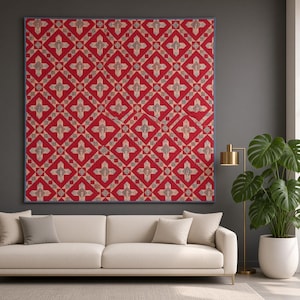 Könnte beinhalten: Ein quadratischer Wandquilt mit einem sich wiederholenden geometrischen Muster aus roten und beigen kreuzförmigen Motiven. Der Quilt hat einen dünnen blauen Rand und hängt an einer dunkelgrauen Wand über einem langen, hellen Sofa mit Kissen.