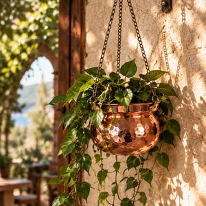 May include: A hammered copper hanging planter with a rounded bowl shape, suspended by three thin metal chains attached to a black iron wall bracket. A lush green trailing plant with heart-shaped leaves grows from the metallic container.