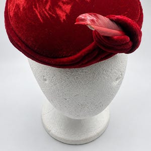 Pode incluir: Um chapéu de veludo vermelho profundo com uma copa arredondada e aba estruturada. O chapéu é adornado com uma única pena vermelha inserida num detalhe de nó de veludo na lateral. Uma pena vermelha a condizer está posicionada abaixo.
