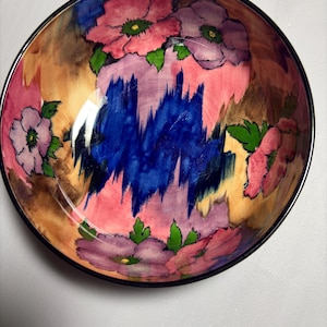 May include: A round ceramic bowl featuring a hand-painted floral pattern with pink and purple flowers, green leaves, and a central abstract splash of deep blue. The background consists of blended tan and brown tones with a dark rim around the edge.