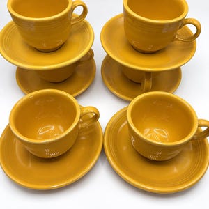 Puede incluir: Un juego de cuatro tazas y platillos de cerámica de color amarillo mostaza a juego. Cada taza presenta una forma de cuenco redondeado con un asa lateral, apoyada sobre un platillo circular con un diseño de anillos concéntricos.