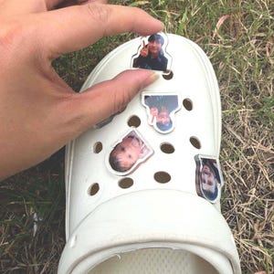 May include: A white foam clog shoe featuring multiple clear acrylic charms attached to the ventilation holes. Each charm displays a unique photographic portrait of a person, with one charm showing a person making a peace sign gesture.