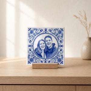Könnte beinhalten: Eine quadratische Keramikfliese mit blau-weißem Blumenmuster am Rand und einem kreisförmigen Porträt eines lächelnden Paares in der Mitte. Die Fliese steht auf einem kleinen, rechteckigen Holzständer und zeigt eine monochrome Illustration.