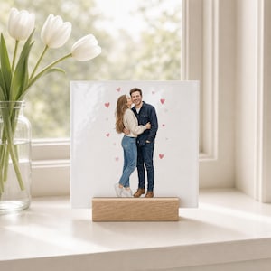 Op de afbeelding: Een vierkante witte keramische tegel met een gedrukte illustratie van een omhelzend stel, omringd door kleine rode hartjes. De tegel wordt ondersteund door een rechthoekige lichtgekleurde houten standaard die als basis dient.