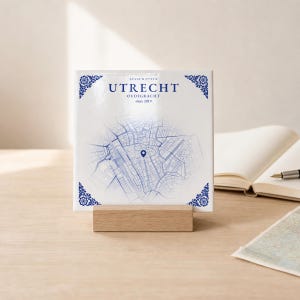 Könnte beinhalten: Eine quadratische weiße Keramikfliese mit einer blauen Karte der Oudegracht in Utrecht, Niederlande. Der Text lautet UTRECHT, OUDEGRACHT und since 2019. Die Fliese hat blaue florale Eckverzierungen und steht auf einem rechteckigen hellen Holzständer.
