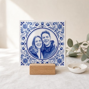 Op de afbeelding: Een vierkante keramische tegel met een blauw-witte bloemenrand en een centraal rond portret van twee personen. De tegel staat op een rechthoekige houten standaard, met daarnaast een klein wit schaaltje met een ring erin.