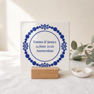 Op de afbeelding: Een vierkante witte keramische tegel met een donkerblauwe cirkelvormige rand van stippen en bloemmotieven. In het midden staat de tekst Emma & James, 14 June 2026, Amsterdam in een donkerblauw lettertype. De tegel staat op een houten standaard.