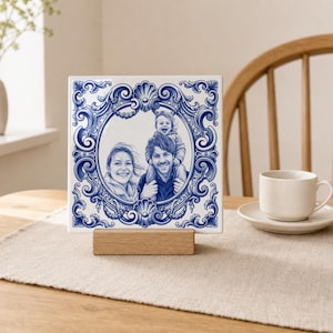 Könnte beinhalten: Eine quadratische Keramikfliese mit einem blau-weißen Zierrand aus komplexen Schnörkelmustern. In der Mitte befindet sich ein monochromes Porträt einer Familie mit einem Erwachsenen und einem Kind, auf einem rechteckigen Holzständer.