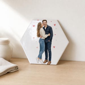 Może przedstawiać: Sześciokątna biała płytka ceramiczna z nadrukowaną ilustracją przedstawiającą dwie przytulające się osoby. Jedna osoba ma na sobie beżowy sweter i niebieskie dżinsy z białymi trampkami, a druga ciemnoniebieską koszulę i dżinsy z brązowymi butami, w otoczeniu małych czerwonych serc.