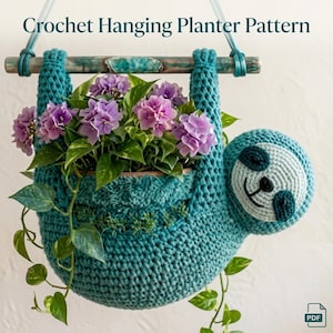Puede incluir: Una maceta colgante de ganchillo color verde azulado con forma de perezoso, que presenta una cara redonda con parches oscuros en los ojos y una boca sonriente. Contiene una planta con flores de hortensia moradas y cuelga de una vara.