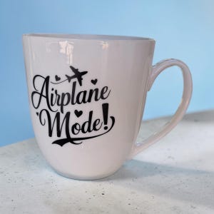 May include: A white ceramic mug featuring black text that reads Airplane Mode! with a small airplane icon and heart shapes. The mug has a curved body and a side handle, presenting a clean design with bold, dark lettering against the smooth white surface.