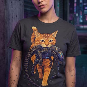 Pode incluir: Uma t-shirt cinzenta escura de manga curta com uma ilustração gráfica de um gato malhado laranja a carregar uma criatura biomecânica escura na boca. O gato tem padrões em espiral na testa e a criatura tem uma cabeça translúcida.