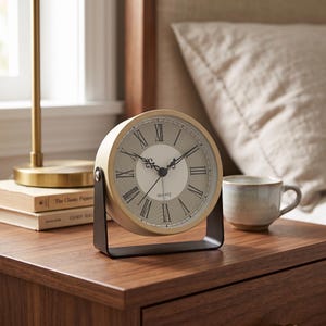 May include: A round quartz table clock with a light-colored frame and a beige face featuring black Roman numerals. The clock is mounted on a dark metal U-shaped stand that allows it to pivot. The word QUARTZ is printed in black on the clock face.