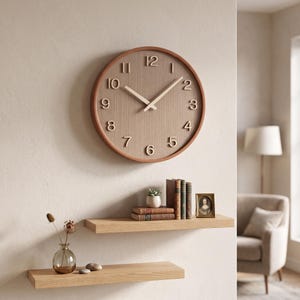 Op de afbeelding: Een ronde wandklok met een bruine rand en een lichtbruine wijzerplaat met houtnerf, voorzien van verhoogde, lichtgekleurde cijfers en bijpassende wijzers. De klok hangt aan een effen muur boven twee houten planken met boeken en decoratie.