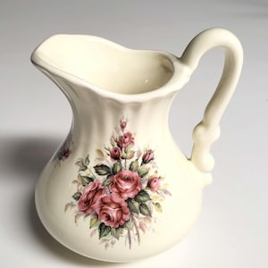 May include: A cream-colored ceramic pitcher featuring a fluted neck and a curved handle. The body of the pitcher is decorated with a floral pattern consisting of pink roses, green leaves, and small buds centered on the front surface.