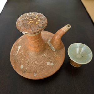 May include: A terracotta ceramic sake server with a wide, flat base, a tall neck, and a curved spout, topped with a textured, gold-flecked lid. Beside the server sits a small, matching ceramic cup with a pale, light blue glazed interior.