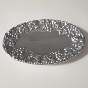 May include: An oval-shaped silver-toned metal tray featuring a raised relief border decorated with detailed grape clusters and vine leaves. The center of the tray has a smooth, reflective surface, while the outer edge is finished with a rope-like texture.