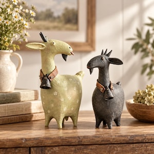 May include: Two goat figurines standing side by side. The larger goat is light green with a white floral pattern, while the smaller goat is dark grey. Both feature textured surfaces, small horns, a goatee, and a brown collar with a metal bell.