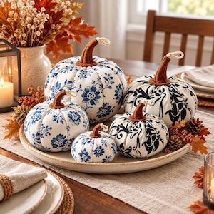 May include: A collection of five fabric pumpkins in varying sizes, featuring white fabric with blue floral patterns or black scrollwork designs. Each pumpkin has a brown felt stem with a small white rope accent attached to the top.