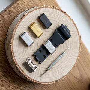 May include: A black watch strap with a silver metal keeper engraved with three stars and the text SINCE 1983. Included are three additional metal keepers in silver, gold, and black, all marked G-SHOCK, a silver buckle, and a metal spring bar tool.