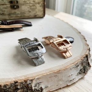 May include: Two metal watch deployment clasps, one in a polished silver tone and one in a rose gold tone, each featuring a prominent T-shaped logo embossed on the front surface. The clasps are designed for watch straps and have a hinged mechanism.