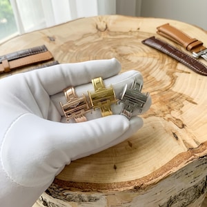May include: Three metal watch deployment clasps in rose gold, yellow gold, and silver tones, each engraved with the Longines logo. The clasps are held together, showcasing their polished finish and mechanical design, with leather watch straps nearby.
