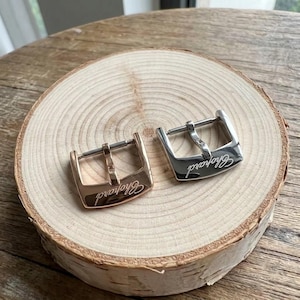 May include: Two metal watch buckles, one in a rose gold tone and one in a silver tone, each featuring the Chopard brand name engraved on the front surface. The buckles have a classic pin design with a rectangular frame and a central prong.