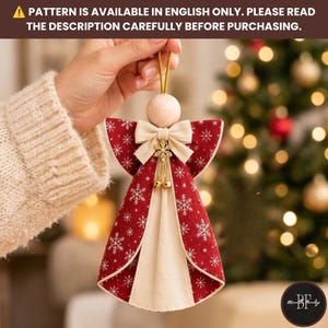May include: A hanging angel ornament featuring a round wooden head, a cream-colored fabric bow, and a red fabric body with a white snowflake pattern. The ornament includes a gold-toned metal flower charm with two small dangling bells.