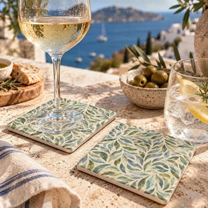 May include: Two square coasters featuring a repeating watercolor pattern of green and blue leaves on a cream background. The coasters are flat with smooth edges and are positioned on a textured surface, one supporting a stemmed wine glass.