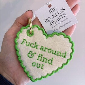 May include: A heart-shaped fabric keyring with a scalloped green embroidered edge and the green embroidered text "Fuck around & find out". It includes a gold-tone metal ring and a white tag labelled "THE RECKLESS HEARTS EMBROIDERED KEEPSAKES".