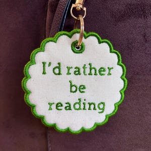 May include: A circular off-white fabric keyring with a scalloped edge and green embroidered border. The text "I'd rather be reading" is embroidered in green thread in the centre. A gold-toned metal lobster clasp and ring are attached.