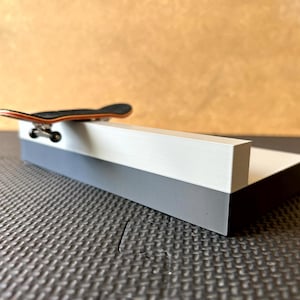 Puede incluir: Un monopatín de dedos en miniatura con tabla negra y bordes laterales naranjas, apoyado sobre un obstáculo rectangular blanco y gris. El borde presenta una superficie superior blanca lisa y una base gris sólida para trucos de fingerboard.