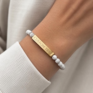 Può includere: Un bracciale composto da perline rotonde in pietra bianca con venature grigie, arricchito da una barra centrale rettangolare in metallo dorato. La barra metallica è incisa con le parole CUSTOM TEXT in un carattere sans-serif, creando contrasto.
