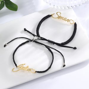 Può includere: Due braccialetti in corda intrecciata nera con targhette in metallo color oro. Un braccialetto riporta il nome Simon e l'altro il nome Julie, entrambi con un piccolo cuore intagliato. Una chiusura magnetica color argento unisce i braccialetti.