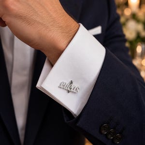 Può includere: Un gemello in metallo color argento con il nome Lucas in carattere corsivo, fissato al polsino bianco di una camicia indossata sotto un blazer blu navy scuro con tre bottoni neri sul polsino della manica.