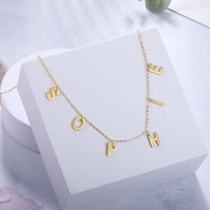 Può includere: Una collana color oro caratterizzata da una catena sottile con ciondoli a forma di lettera appesi singolarmente che compongono il nome SOPHIE. Le lettere sono distanziate lungo la catena, creando un effetto pendente con finitura lucida.